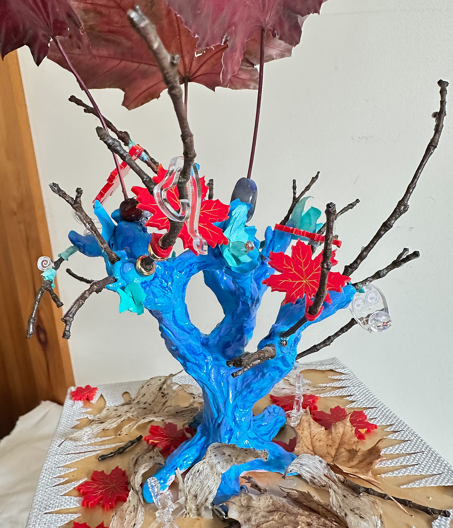 A 3D multi-media piece which depicts a maple wood table on the top grows a blue tree trunk with some bare branches and red leaves. Underneath the table top hang the roots and drops of blood suspended by strings of red beads from the roots. There are words on the droplets like; rapist, enslaver, mother of 12, patriarch, killer, soldier, farmer.